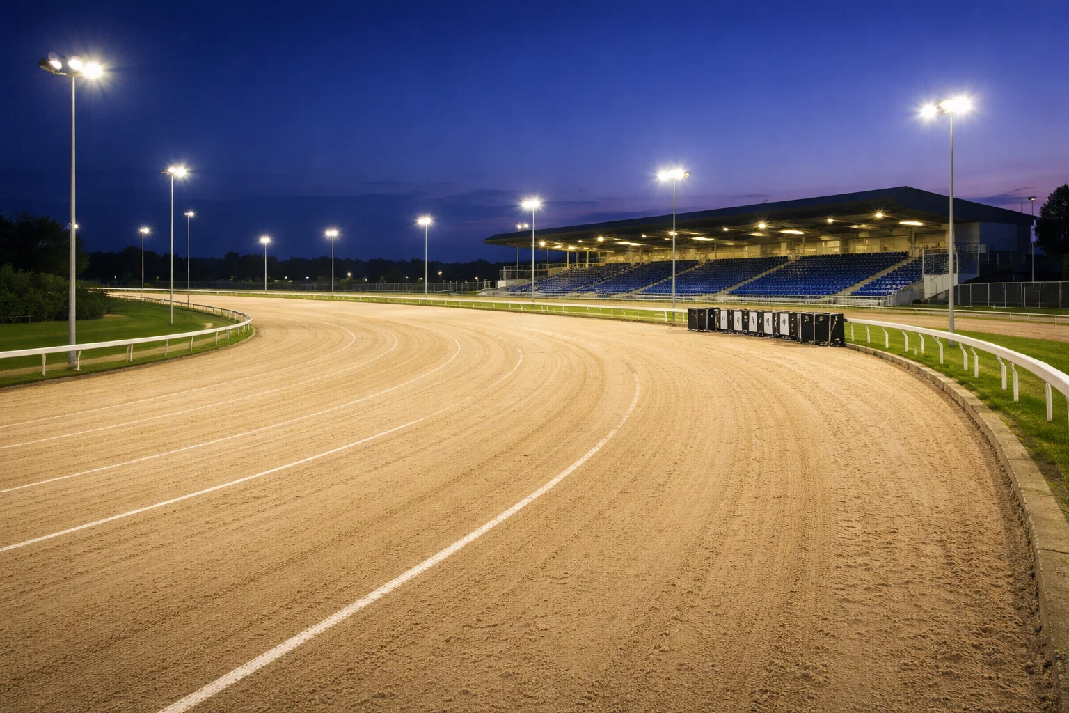 Wide-angle view of a floodlit greyhound racing oval track with sand surface at dusk